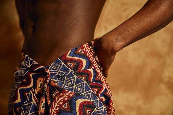 A young handsome African American man confidently poses without a shirt, wearing an eye catching patterned wrap. The warm background enhances his striking presence, capturing a moment of artistry.