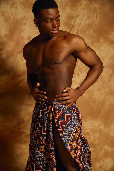 A young, handsome African American man poses confidently without a shirt, adorned in a striking patterned wrap. The warm backdrop highlights his physique and the cultural richness of his outfit.