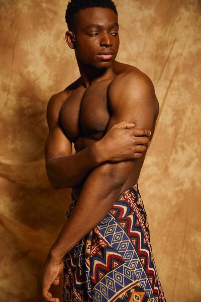 A young, toned African American man stands topless, showcasing his strong physique. Draped in a patterned fabric, he exudes confidence against a warm, artistic backdrop that enhances his presence.