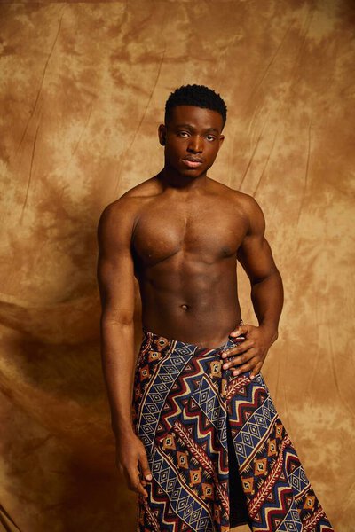 A young confident man stands shirtless, showcasing his toned physique. He wears a patterned wrap that adds a cultural touch. The warm toned backdrop enhances the artistic vibe of the moment.