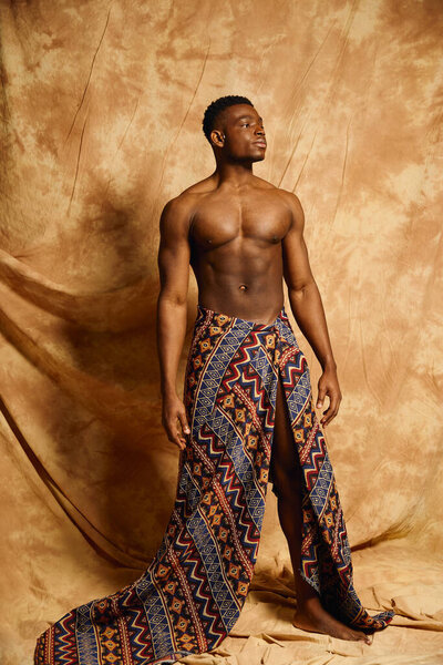 A striking young African American man stands confidently, posing topless in an eye catching patterned garment. The warm backdrop highlights his physique, blending culture and artistry beautifully.