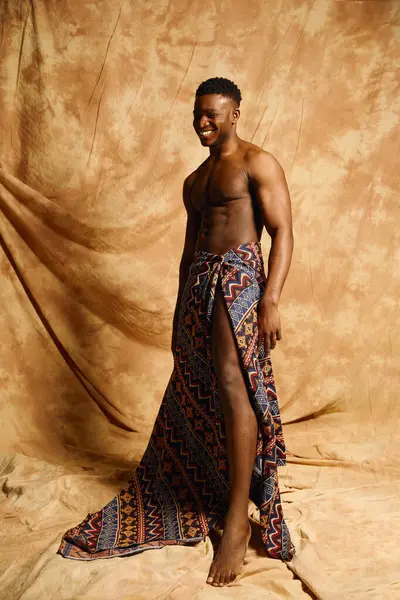 Yakışıklı bir Afro-Amerikan genç adam yaratıcı bir stüdyo ortamında üstsüz poz veriyor. Onun canlı desenli kumaş perdeleri zarif, sıcak bir zemin üzerinde onun fiziğini vurguluyor.