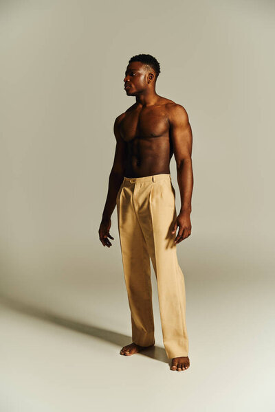 Молодой красивый афроамериканец позирует без верхней одежды в минималистской обстановке. Его уверенная позиция подчеркивает его спортивный телосложение и дополняет моды вперед взгляд с учетом брюки.