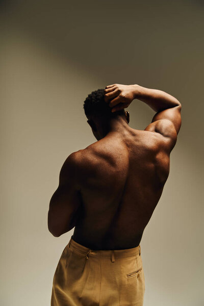 Уверенный в себе молодой человек стоит с сильной осанкой, спиной к зрителю. Он излучает харизму и стиль, подчеркиваемый мягким, художественным освещением, которое усиливает его мышечную форму..