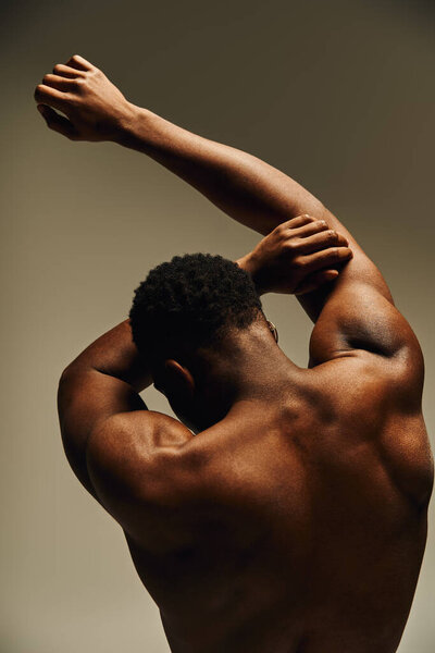 Молодой, красивый афроамериканец уверенно позирует без верхней одежды, демонстрируя свою тонированную физику. Мягкое освещение подчеркивает его черты, создавая художественное ощущение в студии.