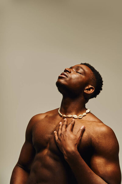 Молодой красивый африканский американец уверенно позирует без рубашки, украшенный уникальным ожерельем. Его выражение выражает чувство силы и спокойствия, создавая художественную атмосферу..