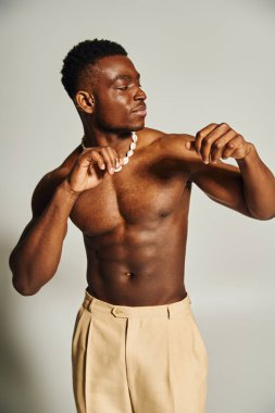 Genç ve yakışıklı bir Afro-Amerikan erkeği, açık renk pantolon giymiş, gömleksiz ve kendinden emin bir şekilde poz verir..