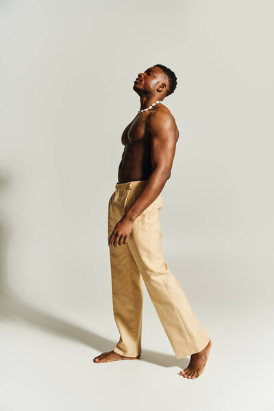 A striking young African American man stands tall and confident, showcasing his physique. He wears cream colored pants and a necklace, embodying a powerful presence in a modern studio.