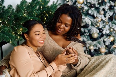 The two women smile affectionately, embraced in warmth and love near a festive tree. clipart