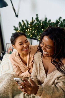 Two women enjoy each others company, laughing and sharing a playful moment near a decorated tree clipart