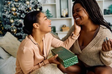 Two women share laughter and warmth while exchanging gifts in a festive holiday setting. clipart