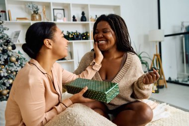 Two women exchange smiles as one surprises the other with a gift on a festive occasion. clipart