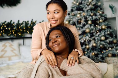 Two women share a warm moment in a festive space adorned with a decorated Christmas tree. clipart