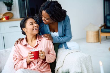 Two women enjoy a joyful morning, sipping tea and embracing love in their cozy home. clipart