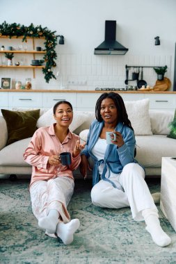 Two women enjoy warm drinks together while sitting on the floor in a cozy living room. clipart