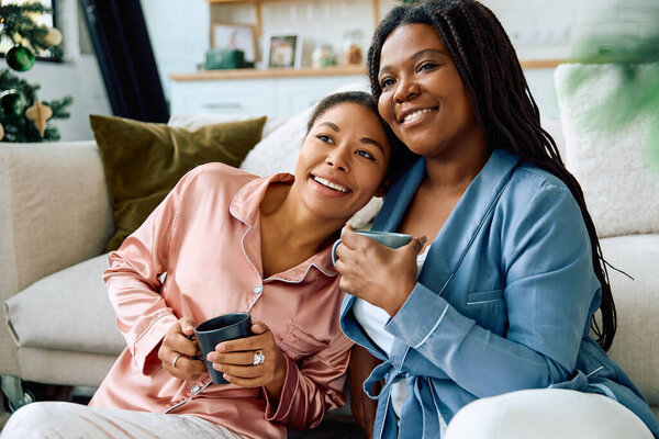 Two women enjoy warm tea together while embracing on a comfy sofa at home.