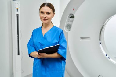 Mavi önlüklü çekici kadın doktor bir hastanede MRI cihazının yanında duruyor..