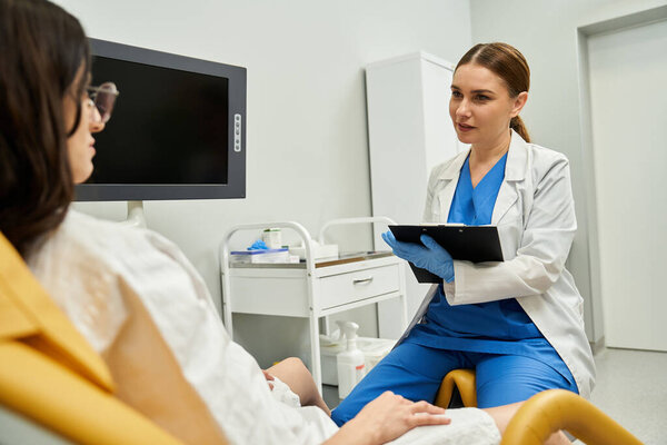 A gynecologist in a lab coat discusses health issues with a female patient during a checkup.