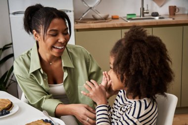 A mother smiles joyfully at her daughter while they enjoy pancakes together in their cozy kitchen. clipart