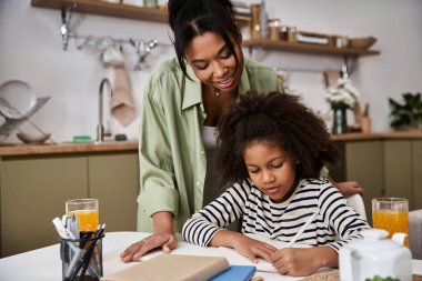 A mother encourages her young daughter as she draws at the kitchen table, sharing warmth and joy. clipart