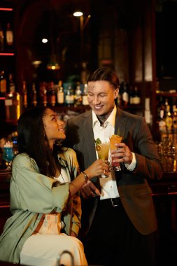 A young couple shares joyful moments while clinking their drinks in a vibrant bar setting. clipart