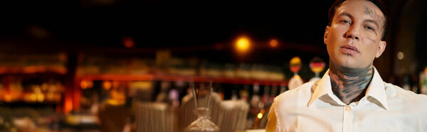 A tattooed young man relaxes in a vibrant bar, surrounded by nightlife energy and dim lights.