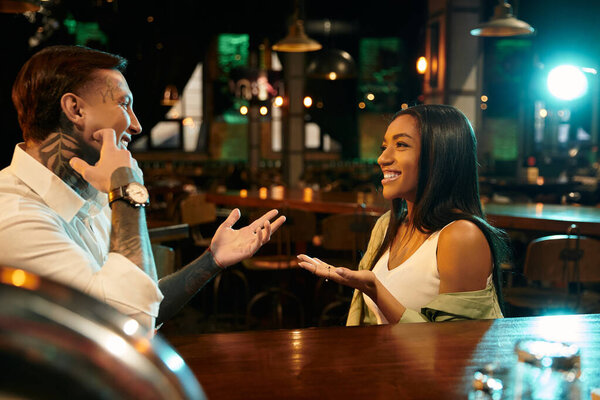 A multicultural couple enjoys their evening date, exchanging smiles and stories in a lively bar.