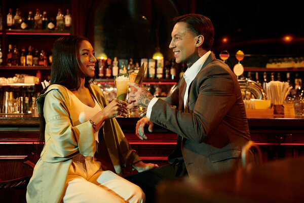 A young couple shares a joyful moment, toasting with cocktails in a lively bar atmosphere.