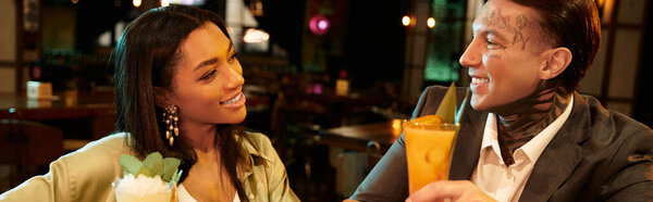 Tattooed young couple shares a joyful moment while sipping cocktails in a vibrant bar.