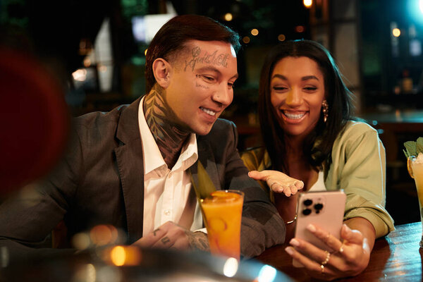 A young couple shares laughter while looking at a phone inside a cozy bar, surrounded by drinks.