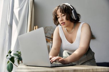 A young woman with curly hair focuses intently on her laptop in a stylish, sunlit room. clipart