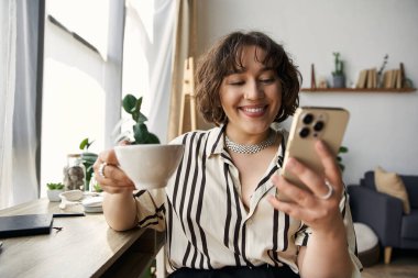 Bright light fills the chic apartment as a young woman smiles, sipping coffee and using her phone. clipart