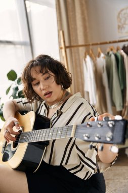 Kıvırcık saçlı genç bir kadın şık dairesinde gitar çalıyor, doğal ışığın tadını çıkarıyor..