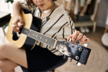 Kıvırcık saçlı genç bir kadın doğal ışıkla dolu şık bir dairede gitarını akort ediyor..
