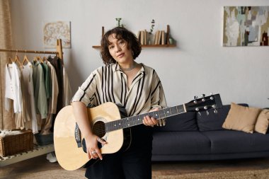 Kıvırcık saçlı genç bir kadın parlak, şık dairesinde gitar çalıyor..