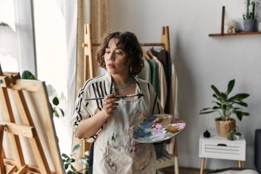 A woman with curly hair paints in her bright, natural-light-filled art space. clipart