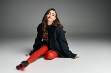 Zarif zarafeti olan genç bir kadın canlı kırmızı taytını ve büyük beden ceketini gösteriyor..