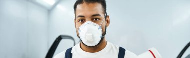 A skilled mechanic wearing a mask and work attire prepares for a task in a busy garage. clipart