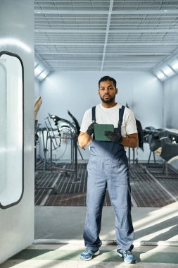 Young mechanic stands focused with clipboard in bright automotive workshop ready for tasks ahead. clipart