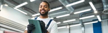 Handsome mechanic smiles while using a tablet, showcasing expertise in a modern auto repair shop. clipart