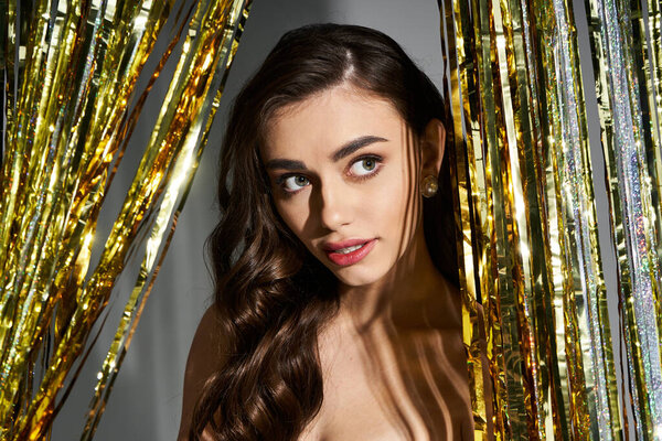 Молодая женщина с текущими волосами смотрит сквозь сверкающие золотые занавески, излучая элегантность.