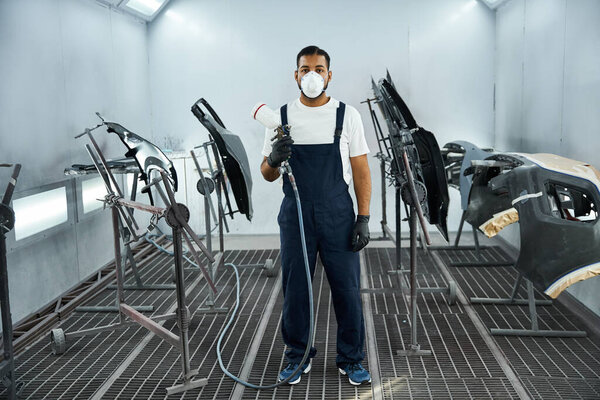 Young mechanic paints car parts in a modern workshop, dressed in safety gear.