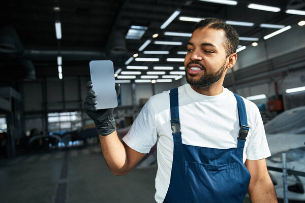 Красивый молодой механик улыбается, показывая свой смартфон в оживленной мастерской..