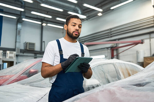 Красивый механик в комбинезоне осматривает машины и пишет важные детали в оживленном гараже.