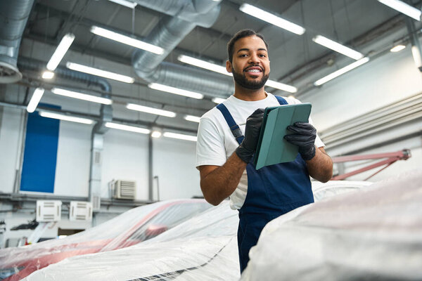 Молодой механик с гордостью стоит с планшетом, проверяя детали автомобиля в занятой мастерской.