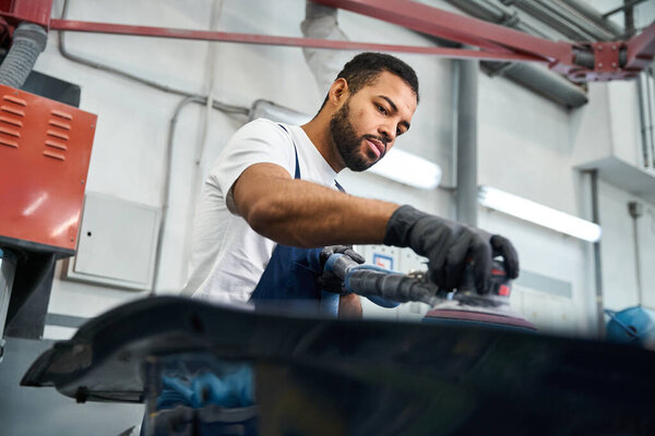 Handsome mechanic uses a power tool to perfect a vehicle part in a busy garage setting.