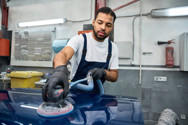 Красивый молодой механик старательно полирует машину в яркой мастерской.