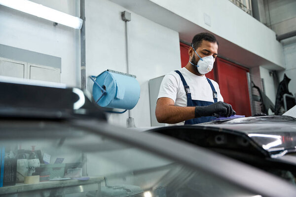 Красивый молодой механик в рабочей одежде прилежно осматривает автомобиль в занятой автомастерской..