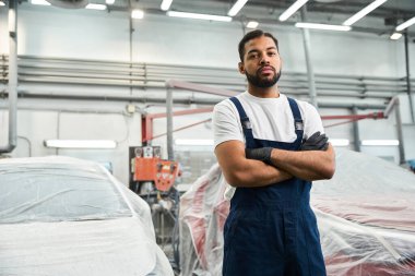 Yakışıklı tamirci kollarını kavuşturmuş, yoğun bir otomotiv dükkanında çalışmaya hazır.