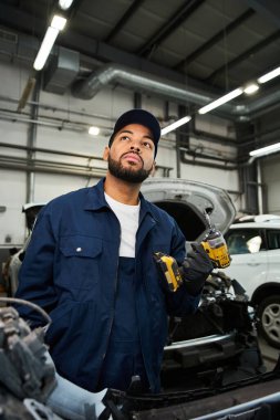 A young mechanic with a thoughtful expression holds tools while working in a bustling garage. clipart